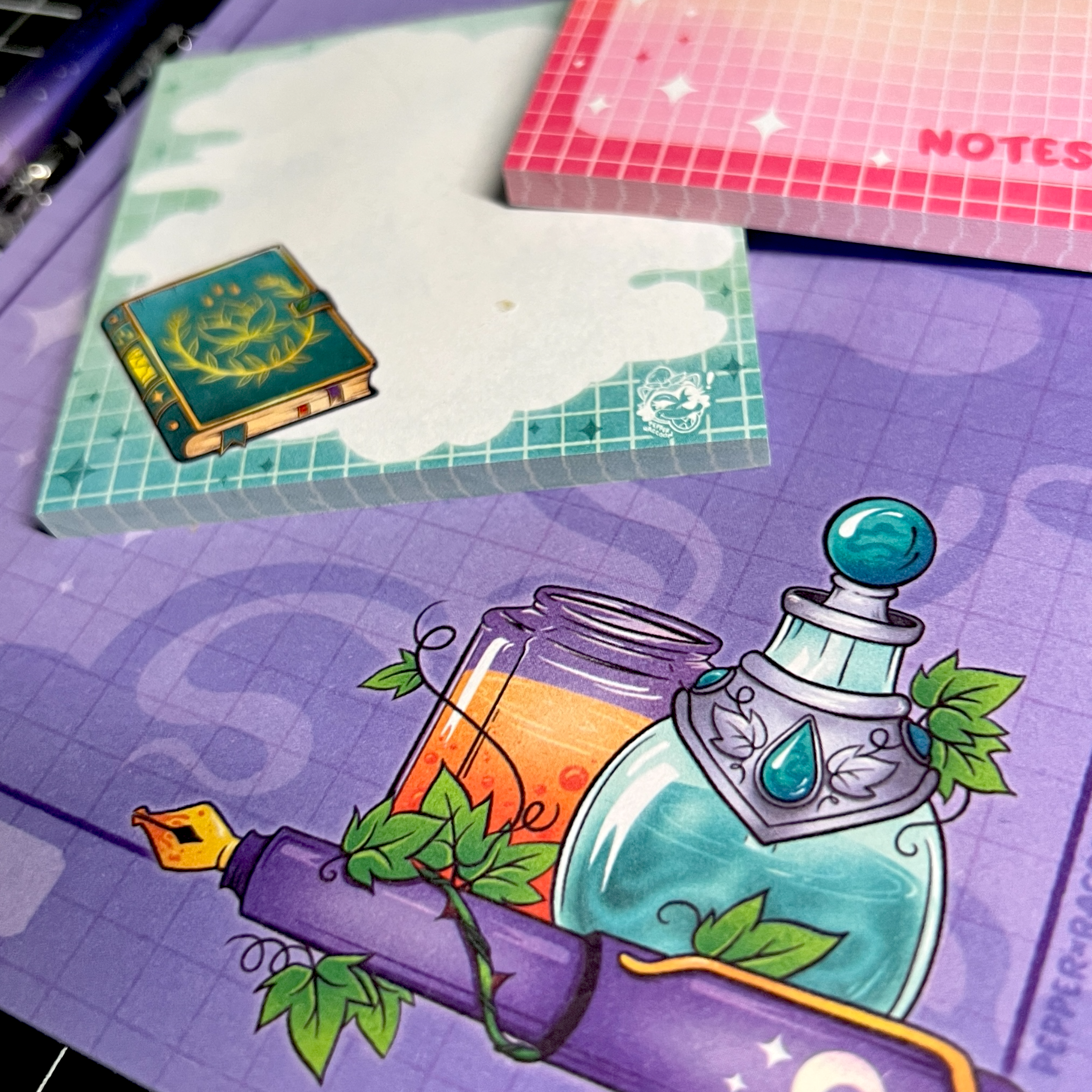 A closeup of a purple notepad with an illustration of several potions and a fountain pen. Atop the notepad are two sticky note pads of different designs.