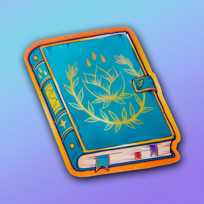 A vinyl sticker of a blue and gold spellbook, featuring plant and rain details, and a latch held shut with a small leaf.