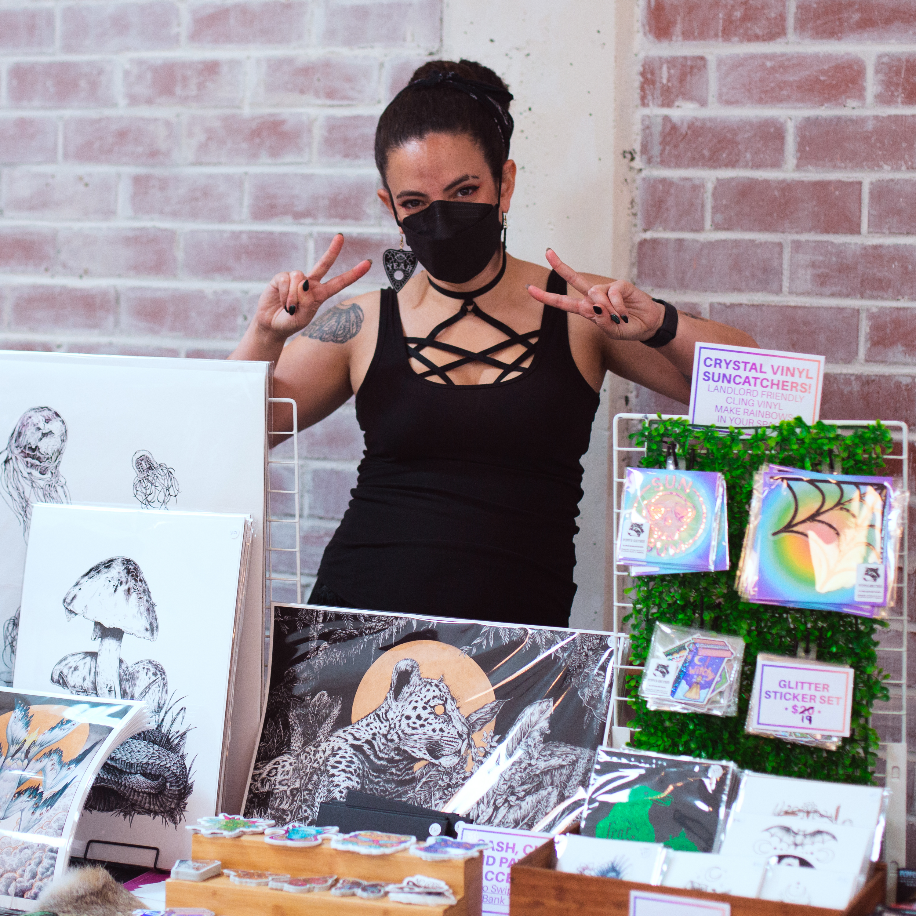 The artist Pepper Raccoon, wearing a face mask and posing behind her artist stall.