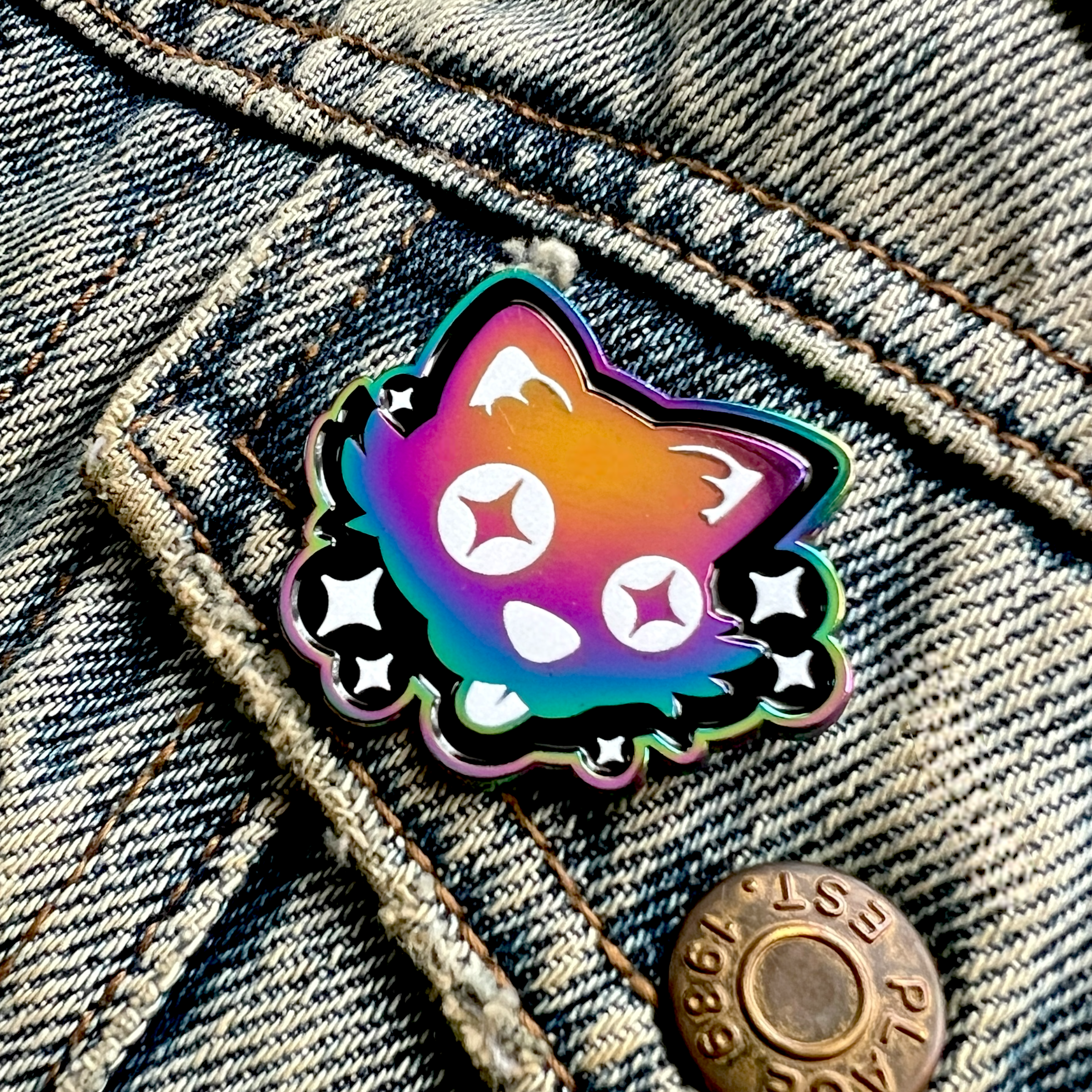 A rainbow anodised enamel pin of a silly cartoon cat, with starry eyes sticking out its tongue. It is surrounded by white sparkles on a black background and is pinned onto a light denim jacket pocket.