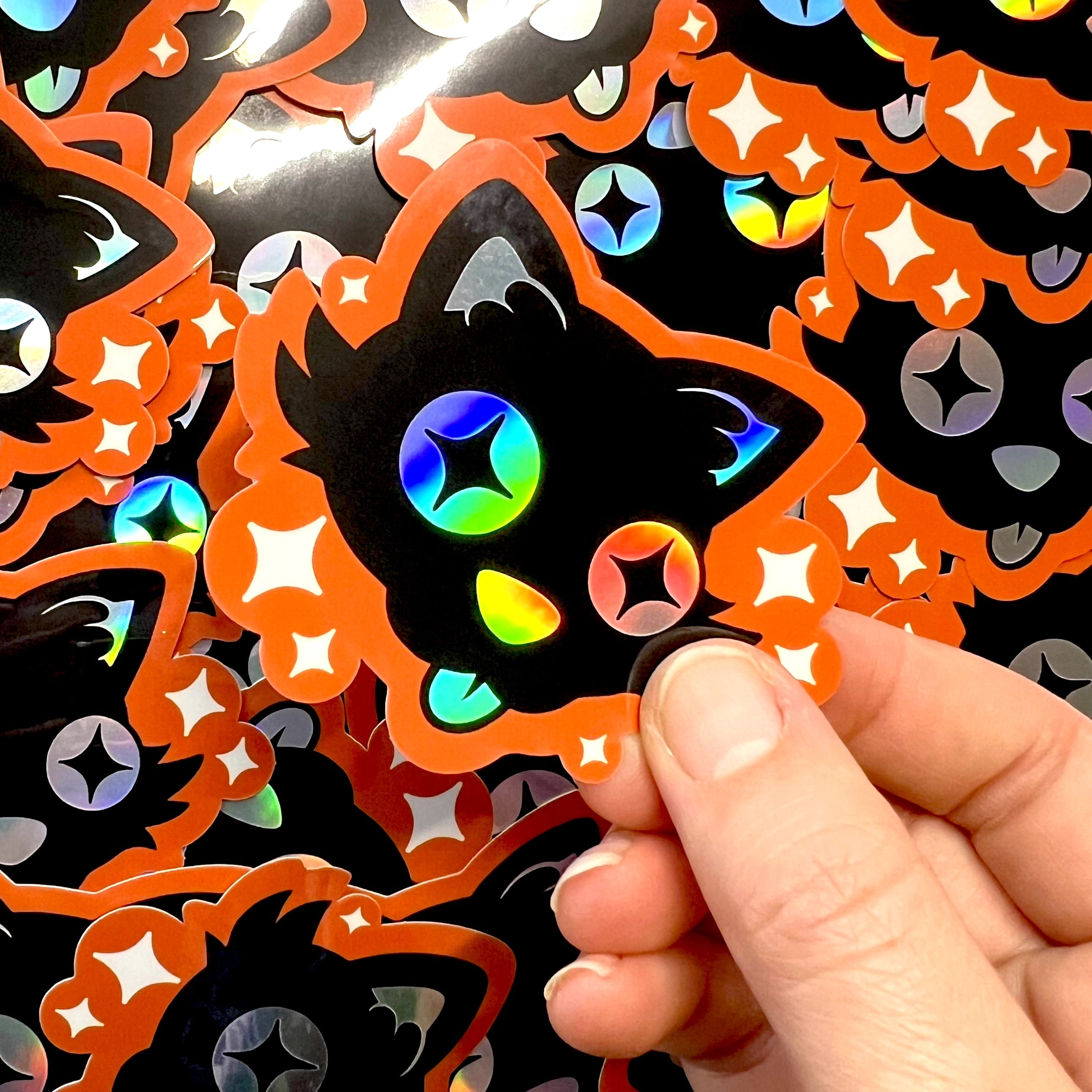 A hand holding an orange and black sticker of a cartoon black cat sticking its tongue out, with white stars and holographic details.