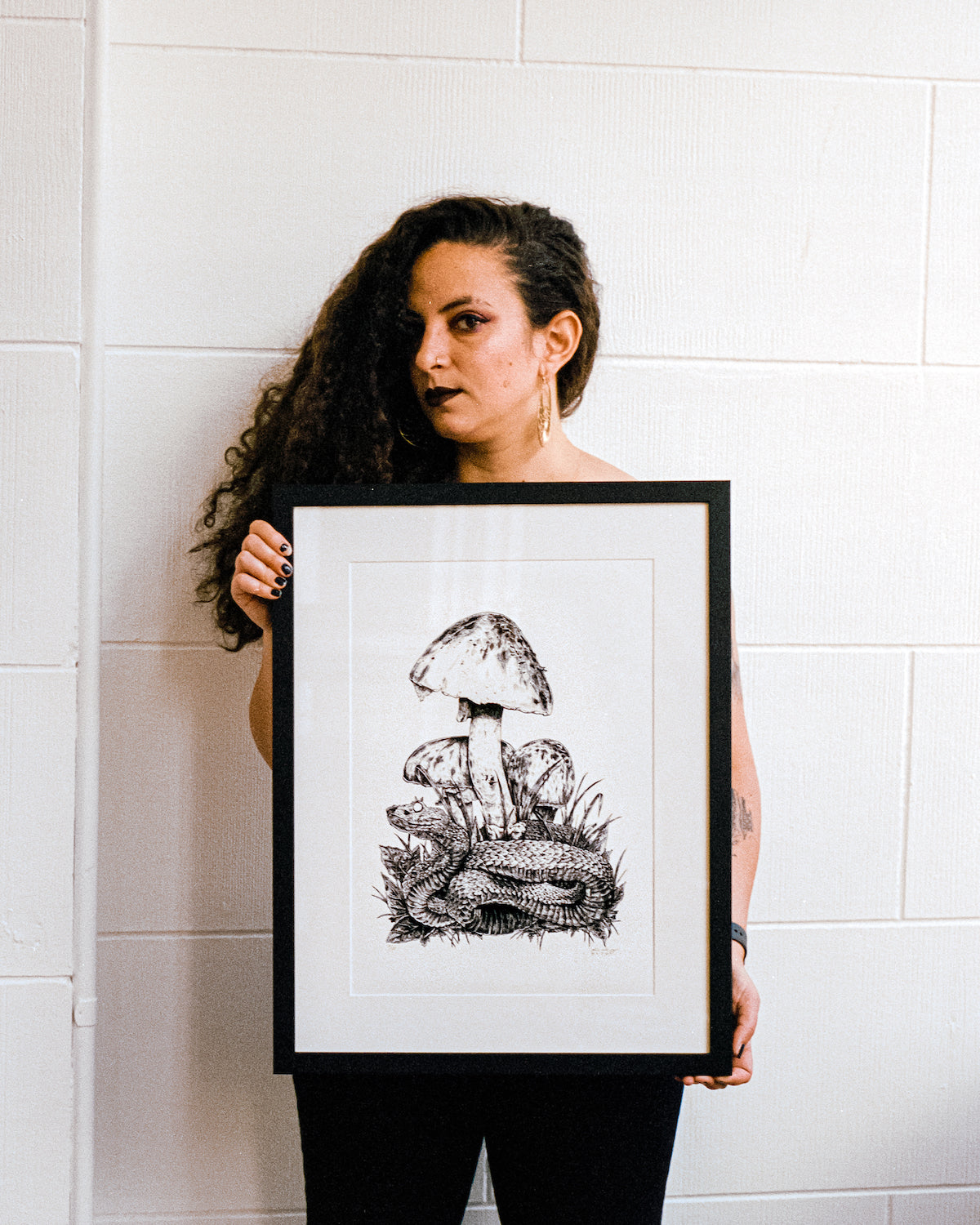 A portrait shot on grainy film of the artist, Pepper Raccoon, holding a large framed ink artwork of an Eyelash Viper.