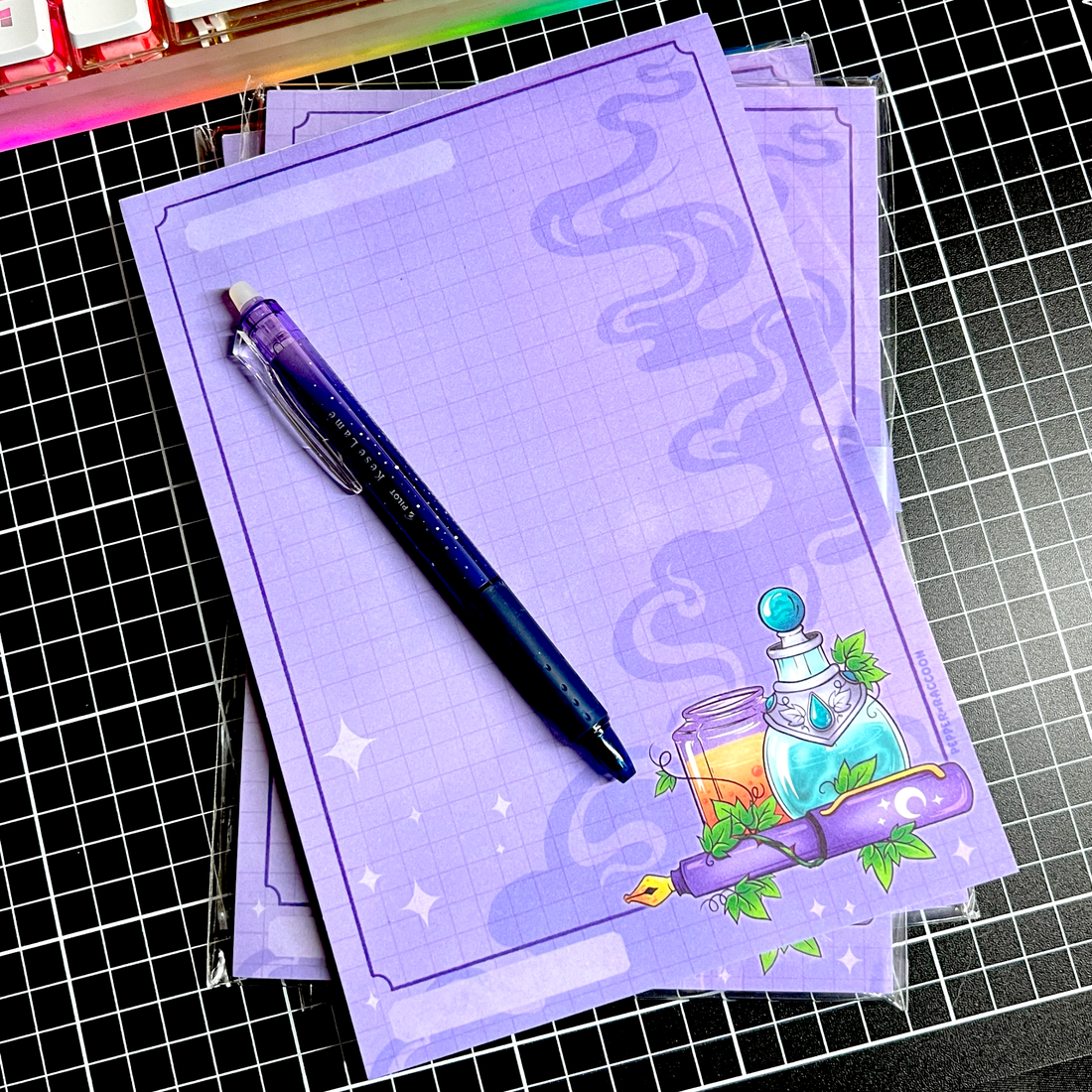 A stack of A5 purple notepads on a black gridded cutting mat. The notepad features purple gridlines, and an illustration of potions and a fountain pen.