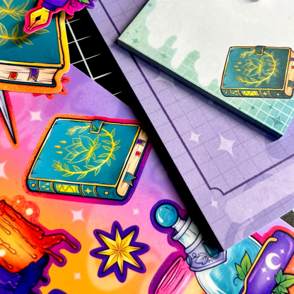 A closeup of a sticker sheet and two notepads featuring Spells + Sparkles witchy artwork of a spellbook, stars, and potions.