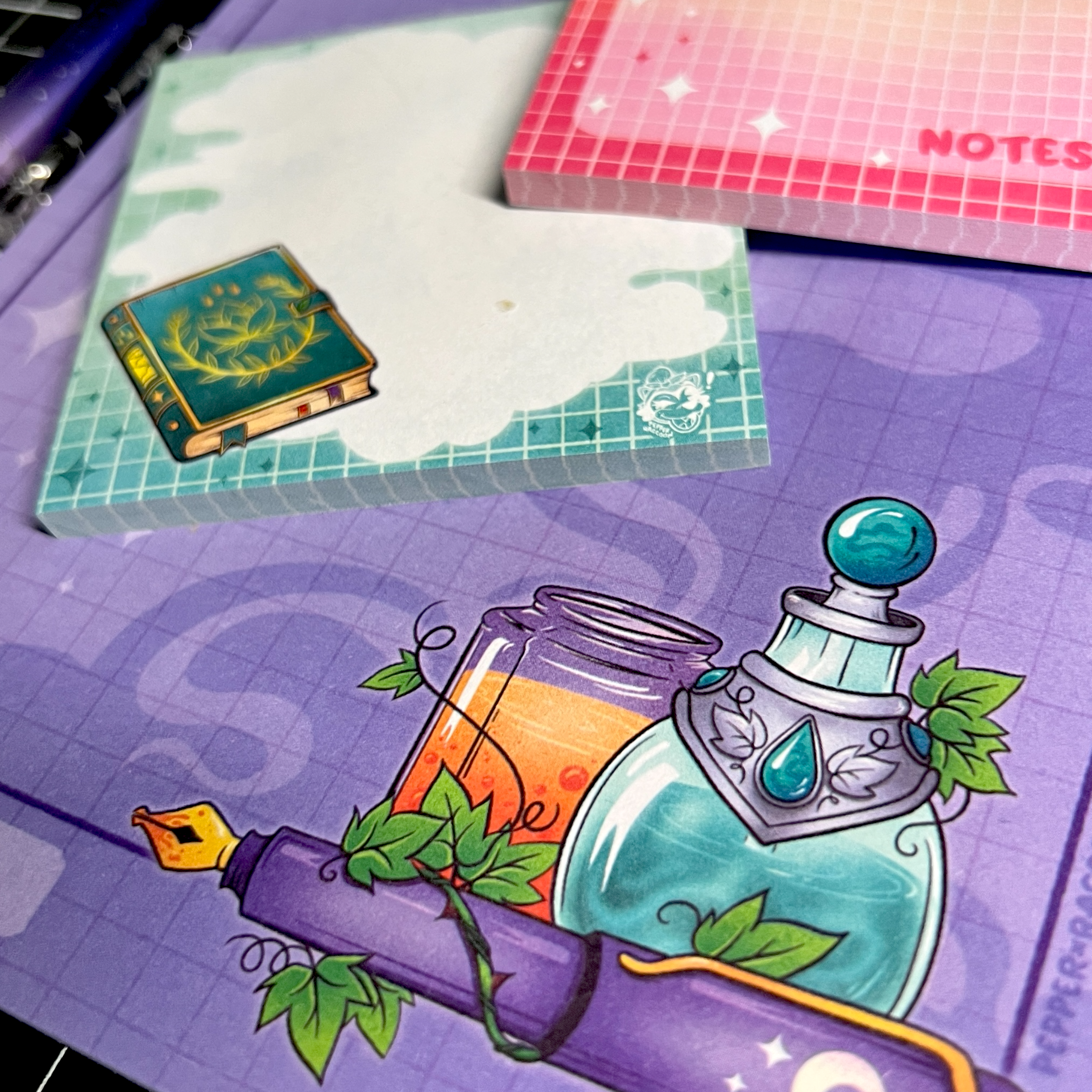 A closeup of a purple notepad with an illustration of several potions and a fountain pen. Atop the notepad are two sticky note pads of different designs. 