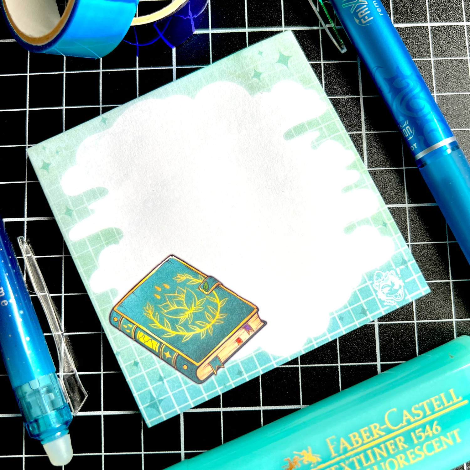 A blue-themed flatlay featuring a sticky note pad with a cloudy area for writing, background grids and fading colours, and an illustration of a spellbook.