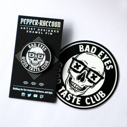 An enamel pin and sticker set featuring a grinning skull wearing nerd glasses and the words Bad Eyes, Good Taste Club.