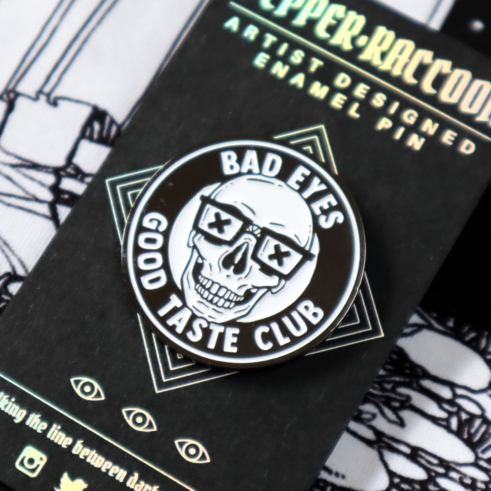 A close-up of an enamel pin featuring a grinning skull wearing nerd glasses and the words Bad Eyes, Good Taste Club.