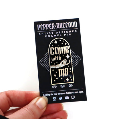 A hand holding an enamel pin on a backing card, the enamel pin says Come With Me in gothic letters with a beckoning skeleton hand.
