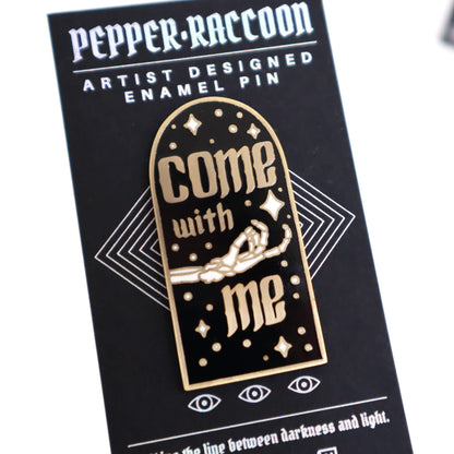 A closer view of an enamel pin that says Come With Me in gothic letters, with a beckoning skeleton hands and stars.